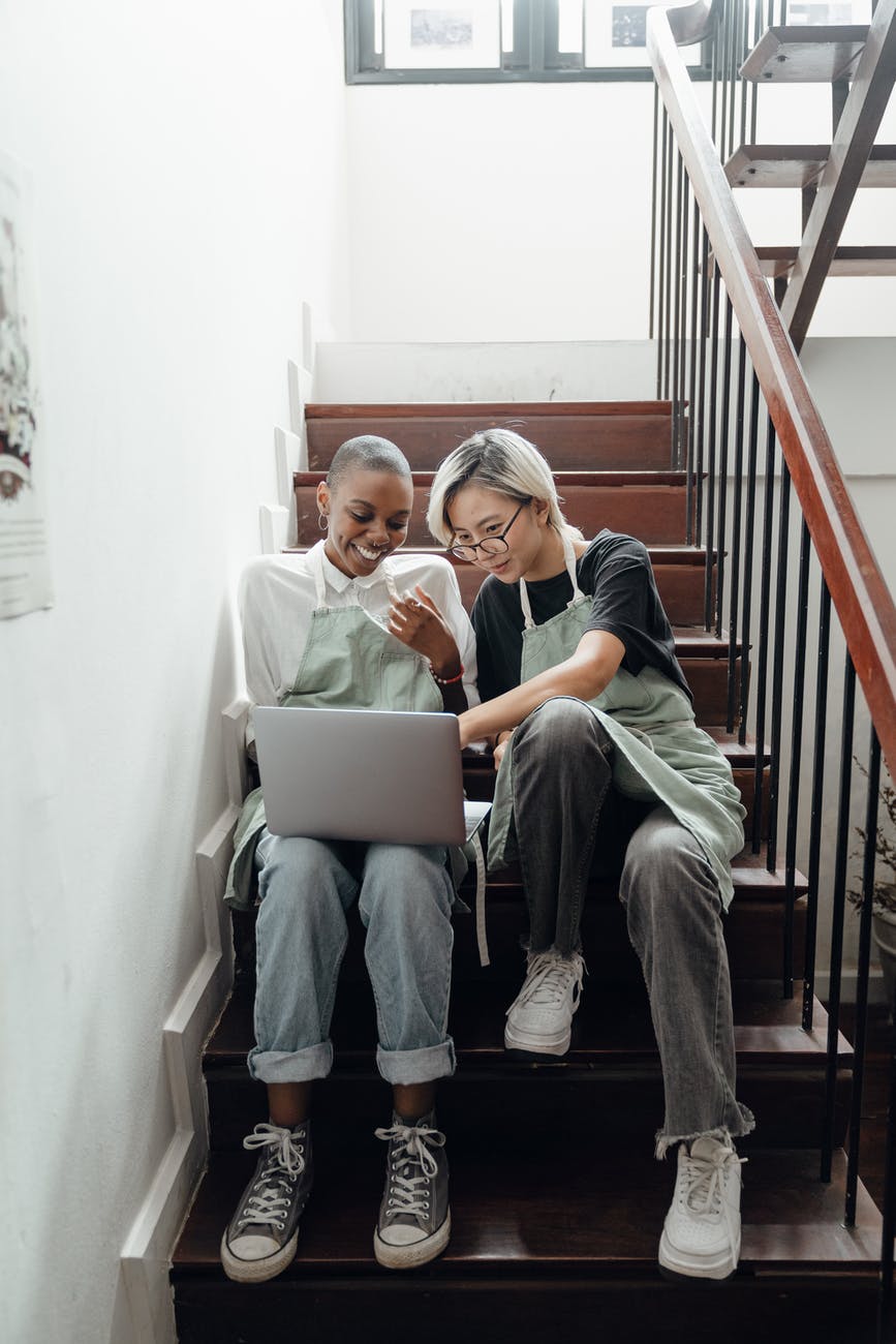happy young multiethnic baristas watching movie on laptop sitting on stairs during break